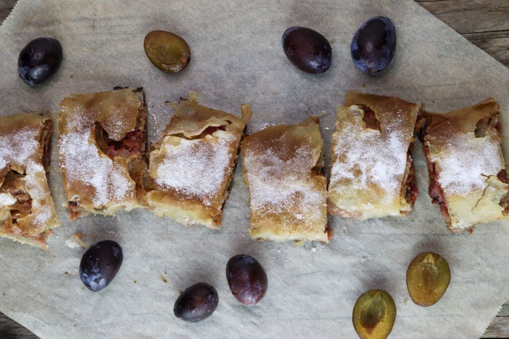 Slices of plum strudel on a piece of parchment paper with fresh plums